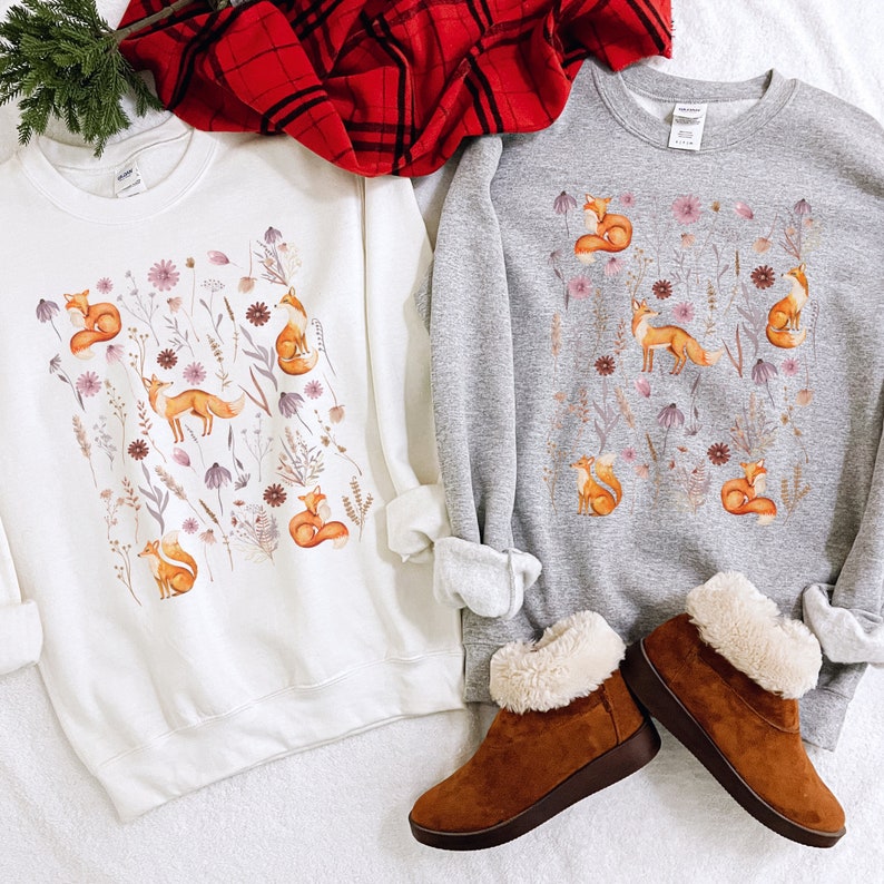 May include: Two sweatshirts with a fox and floral pattern. The sweatshirt on the left is white with a brown fox and purple flowers. The sweatshirt on the right is gray with a brown fox and pink flowers.