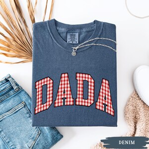 K&ouml;nnte beinhalten: Ein denimblaues T-Shirt mit dem Wort "DADA" in einem rot-wei&szlig; karierten Muster. Eine silberne Halskette mit Anh&auml;nger liegt auf dem Shirt. Das Shirt ist gefaltet, mit einer Jeans und einem wei&szlig;en Schuh sichtbar.