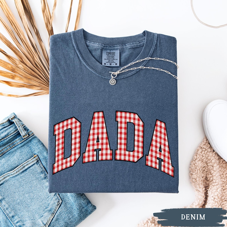K&ouml;nnte beinhalten: Ein denimblaues T-Shirt mit dem Wort "DADA" in einem rot-wei&szlig; karierten Muster. Eine silberne Halskette mit Anh&auml;nger liegt auf dem Shirt. Das Shirt ist gefaltet, mit einer Jeans und einem wei&szlig;en Schuh sichtbar.