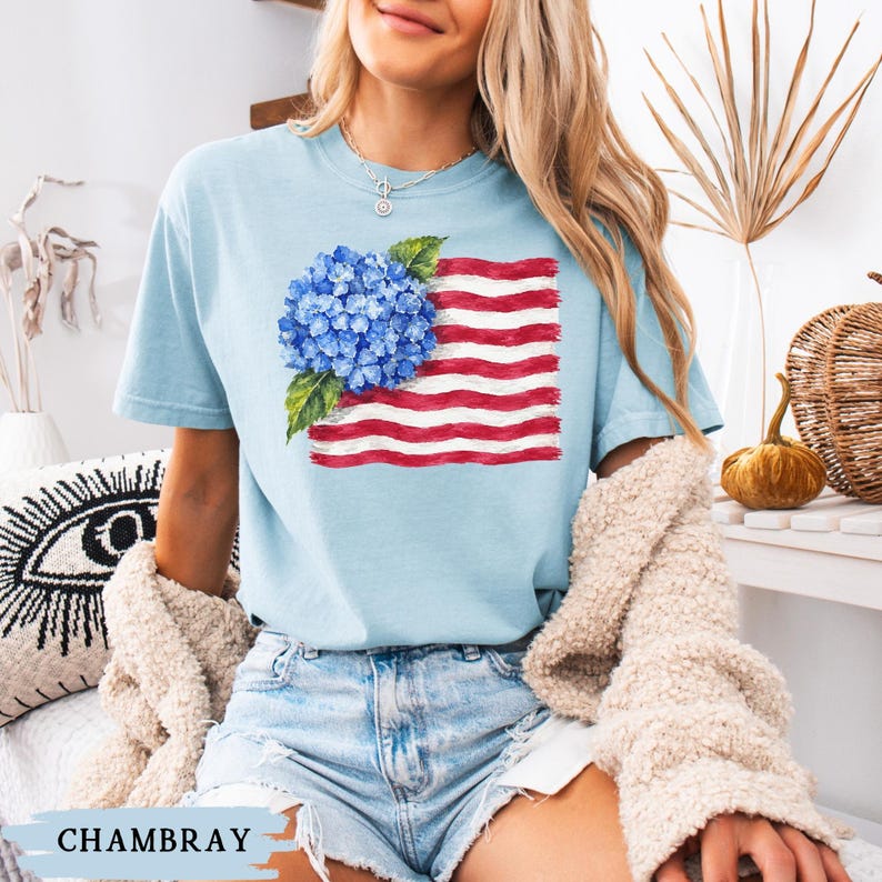 Op de afbeelding: Een lichtblauw t-shirt met korte mouwen en een grafische print van een blauwe hortensiabloem over een gestileerd Amerikaans vlagontwerp met rode en witte strepen. Het shirt heeft een ronde hals en is uitgevoerd in de kleur chambray.
