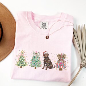 May include: Pink t-shirt featuring a graphic of a chocolate Labrador dog standing in front of three Christmas trees decorated with bows and ornaments. The trees are decorated in different patterns. The text "COMFORT COLORS" is on a white tag attached to the shirt.