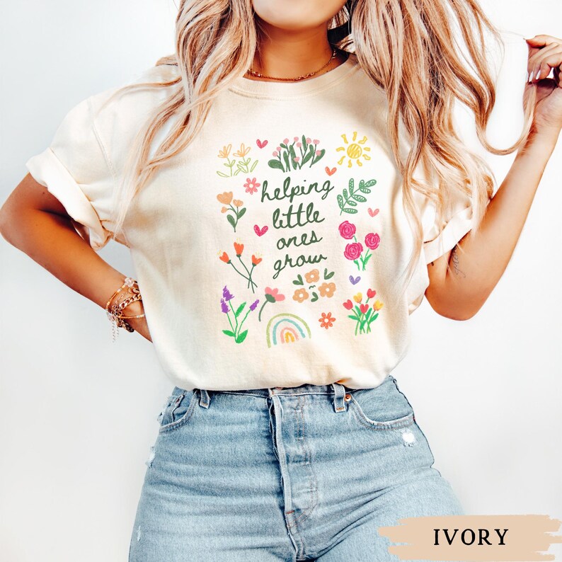 May include: A white t-shirt with a floral design and the text "helping little ones grow". The shirt is being worn by a person with long blonde hair and is paired with blue jeans. The background is white.