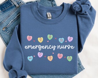 以下が含まれることがあります： 「emergency nurse」という文字がプリントされた青いスウェットシャツ。文字の周りは、小さなキャンディーハートで囲まれており、それぞれ「code love」「love stat」「pulse check」「heart on call」などの言葉が書かれています。