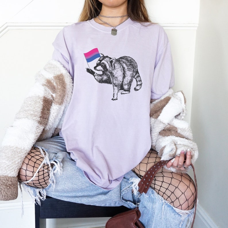 May include: A lavender t-shirt with a graphic of a raccoon holding a bisexual pride flag.