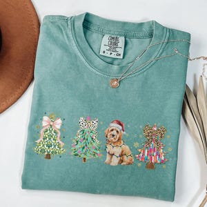 May include: A teal blue t-shirt with a Christmas-themed design featuring three decorated Christmas trees and a dog wearing a Santa hat. The dog is surrounded by Christmas lights. The text "COMFORT COLORS" is visible on the shirt.
