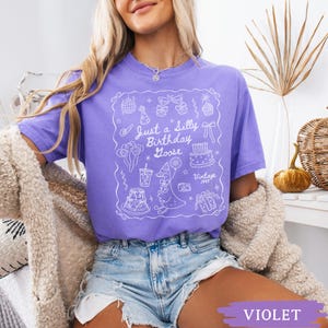 May include: A violet t-shirt with a white graphic that reads "Just a Silly Birthday Goose" with birthday-themed illustrations. The shirt is a short-sleeved, crew neck style. The text "Vintage 1995" is also included in the graphic.