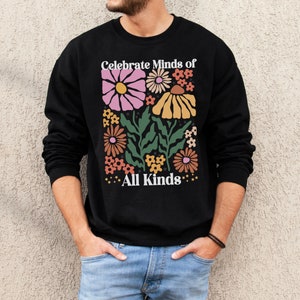 May include: Black sweatshirt with a floral design in pink, yellow, orange, and green. The text "Celebrate Minds of All Kinds" is printed above and below the flowers in white. The sweatshirt is a crew neck style.