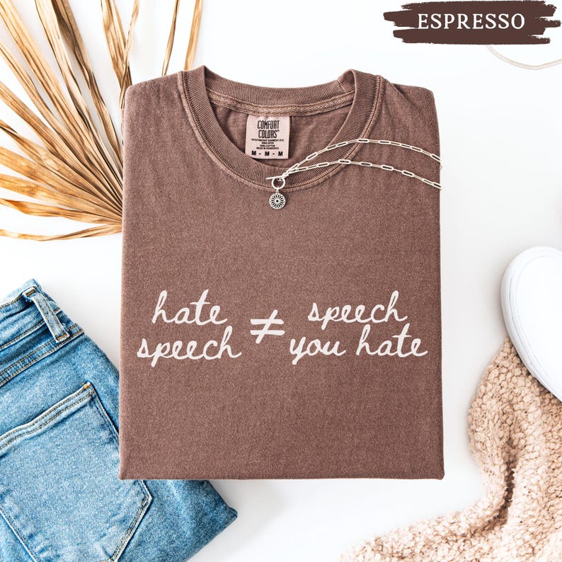May include: Espresso-colored t-shirt with the text "hate speech &ne; speech you hate" in white. The shirt is folded and has a silver necklace with a sun pendant. A pair of blue jeans and a white shoe are visible.