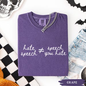 May include: A grape-colored t-shirt with the text "hate speech &ne; speech you hate" in white script. A silver chain necklace is draped over the neckline. The shirt is a Comfort Colors brand.