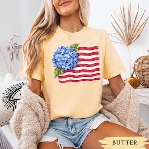 Op de afbeelding: Een lichtgeel t-shirt met korte mouwen voorzien van een grafische print van een blauwe hortensiabloemcluster, geplaatst over een gestileerde Amerikaanse vlag in penseelstreekstijl met rode en witte horizontale strepen en groene bladeren.