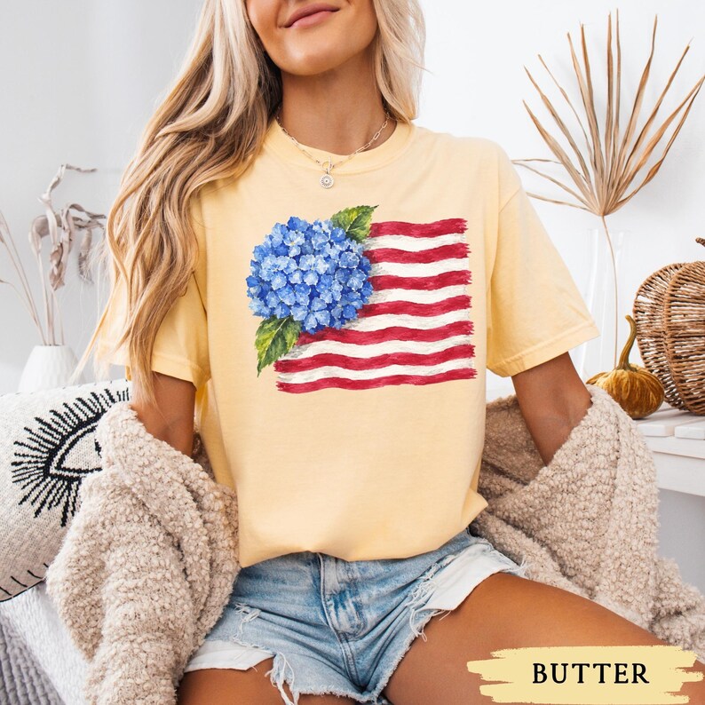 Op de afbeelding: Een lichtgeel t-shirt met korte mouwen voorzien van een grafische print van een blauwe hortensiabloemcluster, geplaatst over een gestileerde Amerikaanse vlag in penseelstreekstijl met rode en witte horizontale strepen en groene bladeren.
