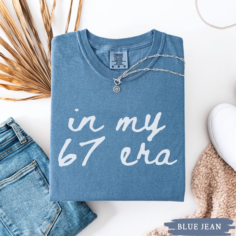 May include: A blue t-shirt with the text "in my 67 era" in white. The shirt is folded and has a silver necklace draped over it. A pair of blue jeans and a white shoe are also in the image.