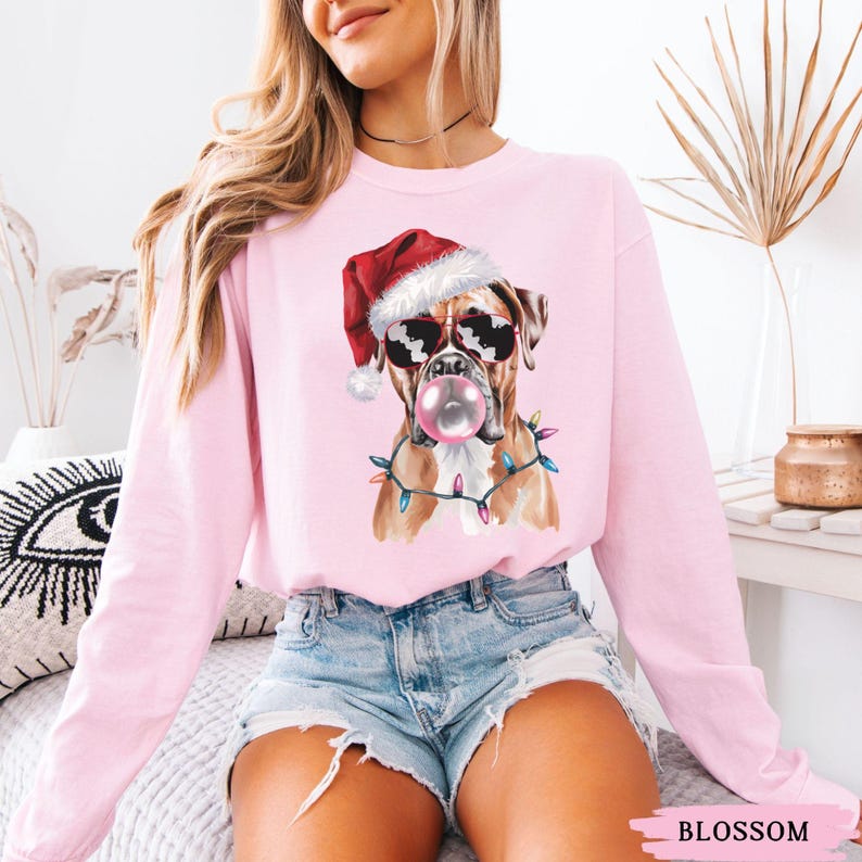 May include: A light pink sweatshirt featuring a Boxer dog wearing sunglasses, a Santa hat, and a string of Christmas lights. The dog is blowing a bubblegum bubble. The word "BLOSSOM" is at the bottom of the image.