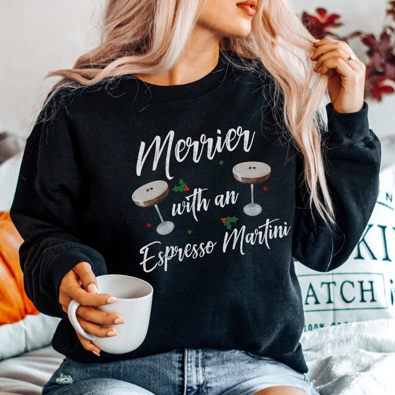 May include: Black sweatshirt with a white graphic that says "Merrier with an Espresso Martini" and features two martini glasses with holly and berries.