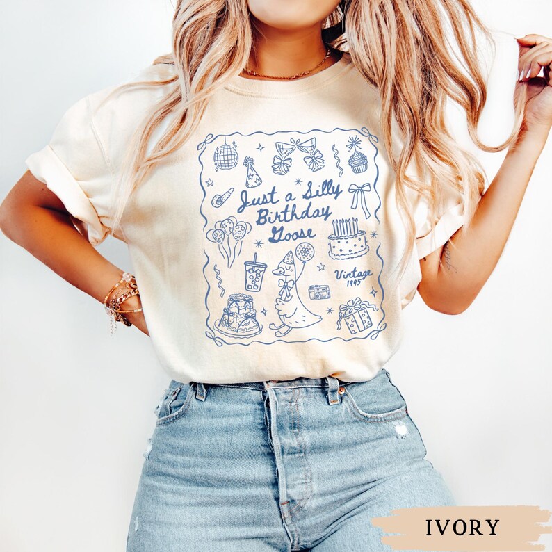 May include: Ivory-colored t-shirt with a blue outlined graphic that reads "Just a Silly Birthday Goose." The graphic includes a goose, cake, balloons, and other birthday-themed illustrations. The text "Vintage 1995" is also included.