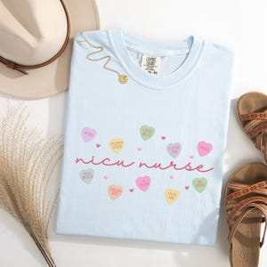 May include: Light blue t-shirt with the text "nicu nurse" in pink script. The shirt is decorated with small pink hearts and candy hearts with various sayings, including "XOXO", "Puppy Love", "Love Bug", "Cutie Pie", "Smile", "You Rock", "Dream Big", "Ever", "Hug Me", and "Be Mine".