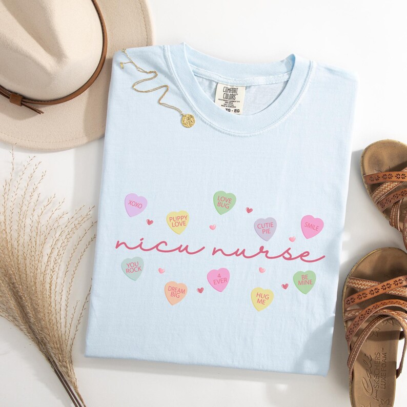 May include: Light blue t-shirt with the text "nicu nurse" in pink script. The shirt is decorated with small pink hearts and candy hearts with various sayings, including "XOXO", "Puppy Love", "Love Bug", "Cutie Pie", "Smile", "You Rock", "Dream Big", "Ever", "Hug Me", and "Be Mine".