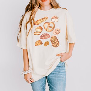 May include: A light beige t-shirt with a graphic print of various baked goods, including bread, pretzels, croissants, and donuts.
