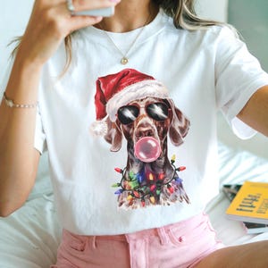 May include: White t-shirt featuring a brown and white dog wearing sunglasses and a Santa hat, blowing a bubblegum bubble. The dog is adorned with a string of colorful Christmas lights. A fun, festive design.