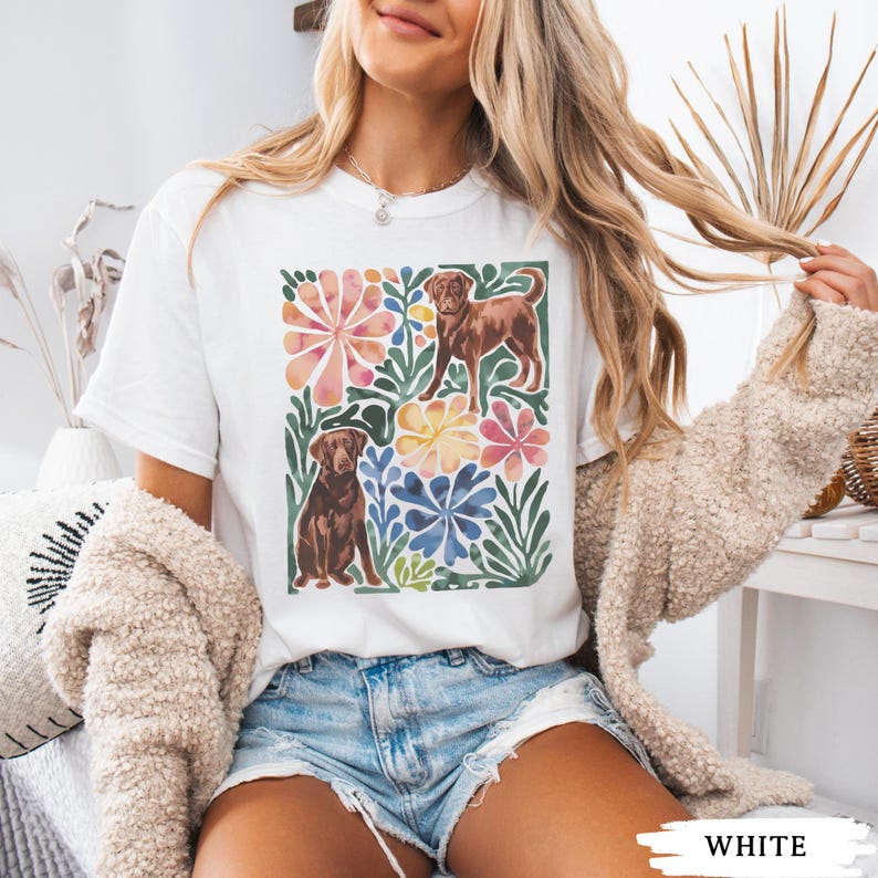 May include: White t-shirt with a watercolour design of two brown dogs surrounded by colourful flowers and green leaves. The design includes pink, yellow, and blue floral elements. The word "WHITE" is printed at the bottom.