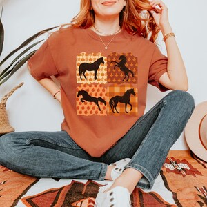 May include: A rust-colored t-shirt with a graphic of four black silhouettes of horses in a patchwork design. The design features a variety of fall-themed patterns, including plaid, leaves, and pumpkins.