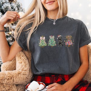 May include: A dark gray t-shirt with a Christmas tree design featuring a black dog, a leopard print tree, a green and gold tree, and a colorful tree with a bow.