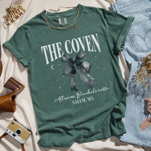 K&ouml;nnte beinhalten: Ein gr&uuml;nes T-Shirt mit einer wei&szlig;en Grafik, auf der "The Coven" mit einer schwarzen Schleife und Sternen steht. Der Text unter der Schleife lautet "Allison's Bachelorette Salem, MA."