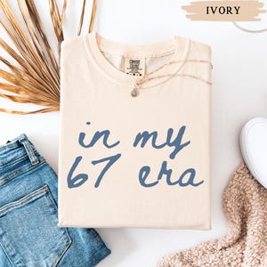 May include: Ivory-colored t-shirt with the text "in my 67 era" in blue script. The shirt is folded and displayed with a gold necklace, blue jeans, and a white shoe. The shirt is made by Comfort Colors.