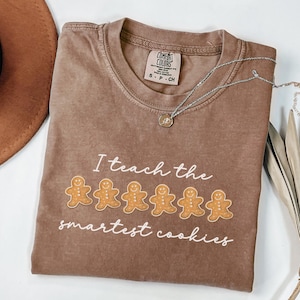 May include: A brown Comfort Colors t-shirt with the text "I teach the smartest cookies" and gingerbread man graphics. A silver necklace with a pendant is draped across the shirt.