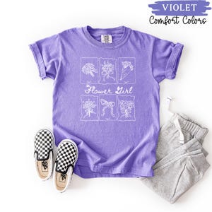 May include: A violet Comfort Colors t-shirt with a white floral design and the words "Flower Girl." The shirt is paired with gray sweatpants and black and white checkered slip-on shoes.