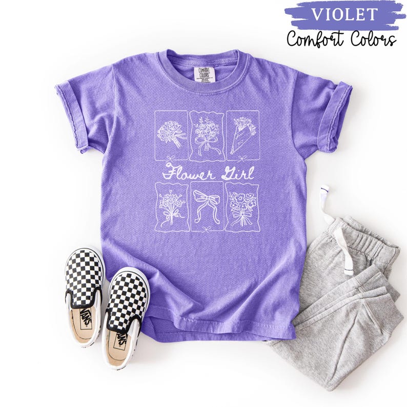 May include: A violet Comfort Colors t-shirt with a white floral design and the words "Flower Girl." The shirt is paired with gray sweatpants and black and white checkered slip-on shoes.
