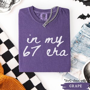 May include: A grape-colored short-sleeve t-shirt with the text "in my 67 era" in white script. A silver chain necklace is draped across the shirt. The shirt is folded on a white surface.