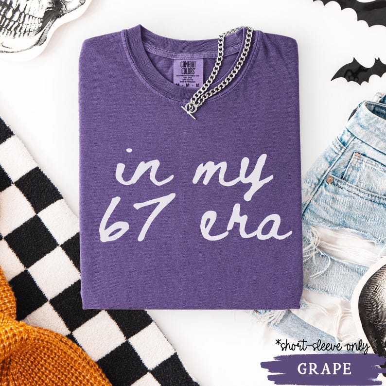 May include: A grape-colored short-sleeve t-shirt with the text "in my 67 era" in white script. A silver chain necklace is draped across the shirt. The shirt is folded on a white surface.