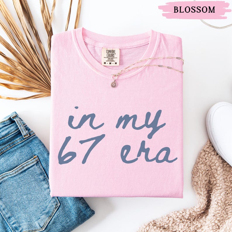 May include: A light pink t-shirt with the text "in my 67 era" in blue. A gold necklace with a pendant rests on the shirt. The shirt is folded and placed on a white surface, next to a pair of blue jeans and a white shoe.