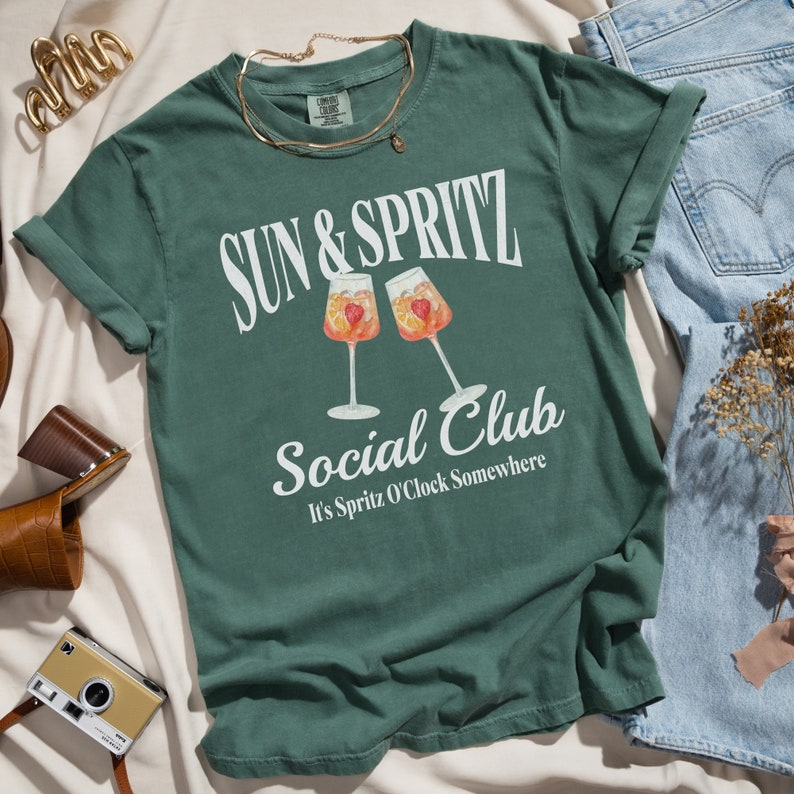May include: A green t-shirt with the text "Sun & Spritz Social Club" and the phrase "It's Spritz O'Clock Somewhere" printed on it. The shirt features a graphic of two glasses of a cocktail with orange slices and strawberries.