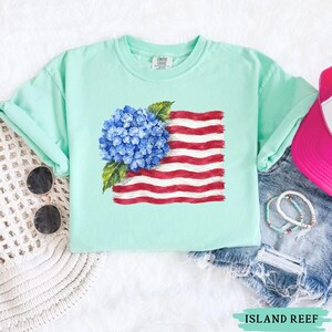 Op de afbeelding: Een licht mintgroen t-shirt met een grafische print van een blauwe hortensiabloem die een gestileerd vlagontwerp met rode en witte strepen overlapt. Het shirt heeft een ronde hals en korte mouwen voor een casual en ontspannen uitstraling.