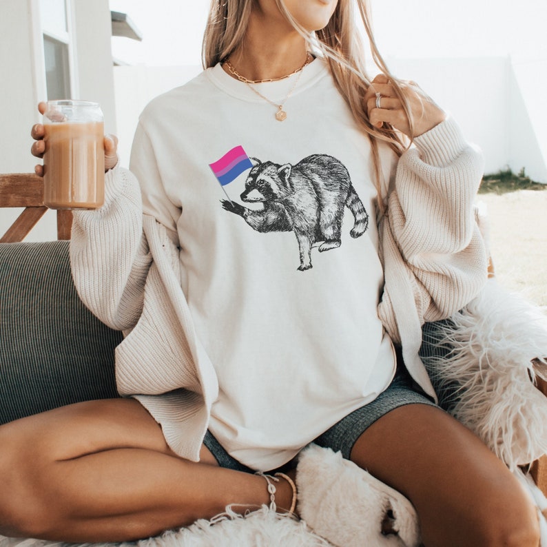 May include: A white crew neck sweatshirt with a graphic of a raccoon holding a bisexual pride flag. The raccoon is black and white and the flag is blue, pink, and purple.