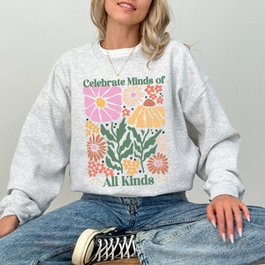 May include: A light gray sweatshirt with a floral design and the text "Celebrate Minds of All Kinds." The graphic features pink, orange, and brown flowers with green leaves. The sweatshirt is a crew neck style.