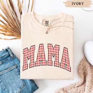 K&ouml;nnte beinhalten: Elfenbeinfarbenes Sweatshirt mit dem Wort "MAMA" in gro&szlig;en, rot-wei&szlig; karierten Buchstaben. Das Shirt ist gefaltet und liegt auf einer wei&szlig;en Oberfl&auml;che neben einer Jeans und einem wei&szlig;en Schuh.