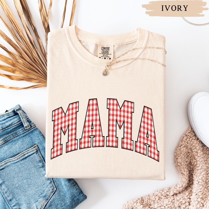 K&ouml;nnte beinhalten: Elfenbeinfarbenes Sweatshirt mit dem Wort "MAMA" in gro&szlig;en, rot-wei&szlig; karierten Buchstaben. Das Shirt ist gefaltet und liegt auf einer wei&szlig;en Oberfl&auml;che neben einer Jeans und einem wei&szlig;en Schuh.