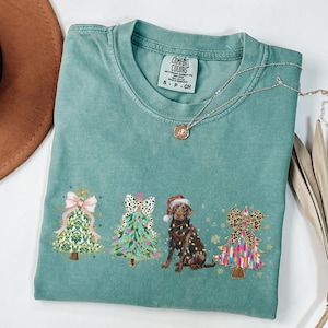 May include: A teal blue t-shirt with a Christmas-themed design featuring three decorated Christmas trees and a chocolate Labrador wearing a Santa hat. The trees are decorated with bows, ornaments, and lights. The text "COMFORT COLORS" is visible on the tag.