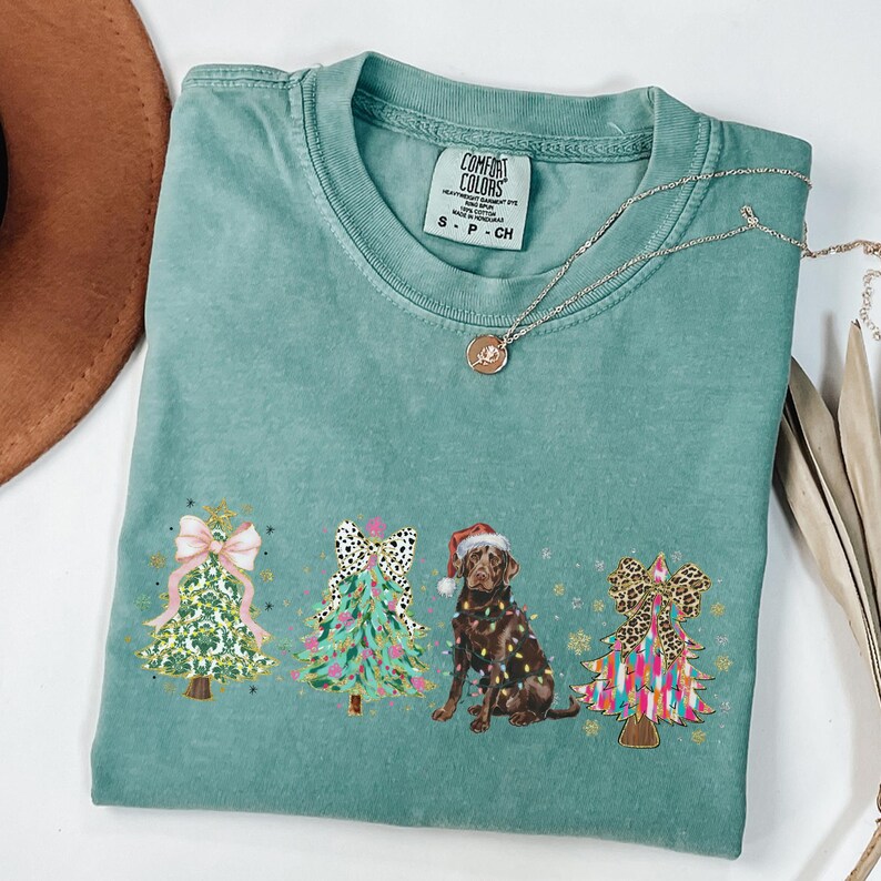 May include: A teal blue t-shirt with a Christmas-themed design featuring three decorated Christmas trees and a chocolate Labrador wearing a Santa hat. The trees are decorated with bows, ornaments, and lights. The text "COMFORT COLORS" is visible on the tag.