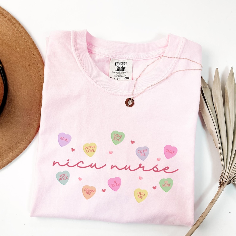 May include: Pink t-shirt with the text "nicu nurse" in a cursive font. The text is surrounded by colorful candy hearts with various sayings, such as "XOXO", "Puppy Love", "Love Bug", "Cutie Pie", "Smile", "You Rock", "Dream Big", "4 Ever", "Hug Me", and "Be Mine".