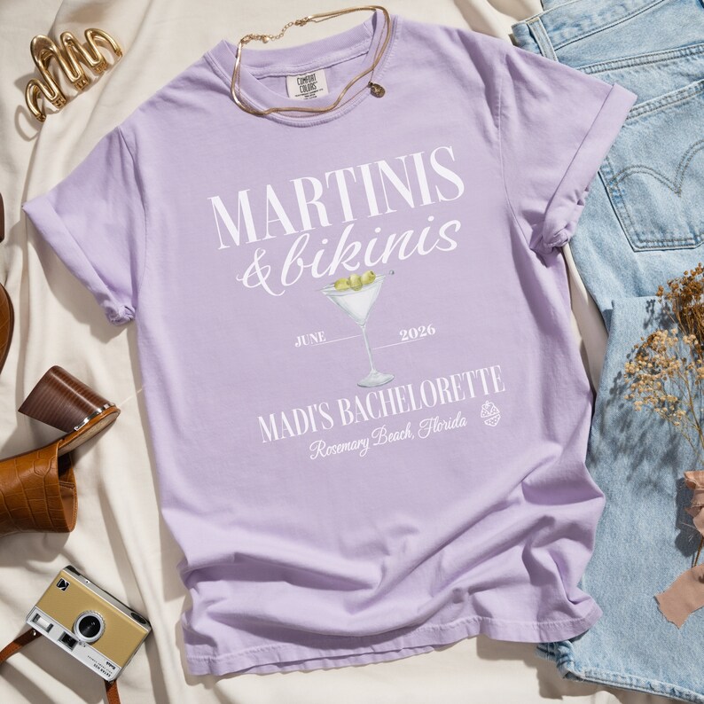 May include: A lavender t-shirt with the text "MARTINI'S & bikinis" in white. The shirt also features a martini glass with olives, the text "JUNE 2026", and "MADI'S BACHELORETTE Rosemary Beach, Florida".