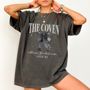 K&ouml;nnte beinhalten: Ein &uuml;berdimensioniertes, dunkelgraues T-Shirt mit einem grafischen Aufdruck eines schwarzen Schleifchens mit einer Mondsichel und Sternen. Der Text "THE COVEN" ist &uuml;ber dem Schleifchen gedruckt und "Allison's Bachelorette SALEM, MA" unter dem Schleifchen.