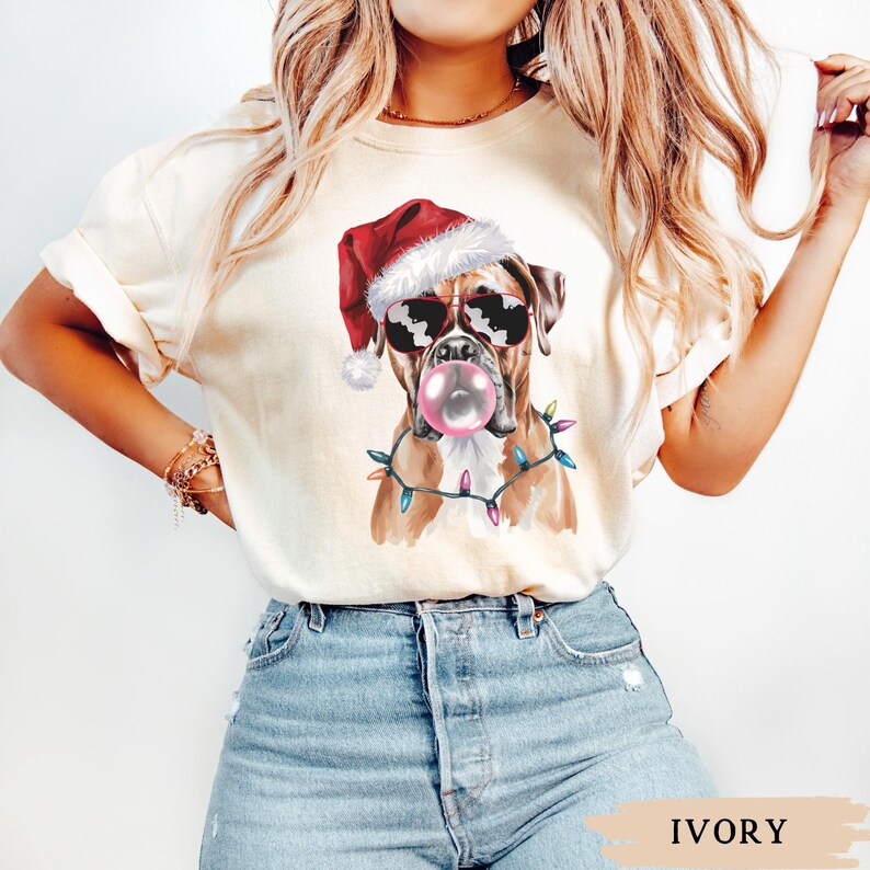 May include: Ivory-colored t-shirt featuring a Boxer dog wearing sunglasses and a Santa hat, blowing a bubblegum bubble. The dog is adorned with a string of colorful Christmas lights. The word "IVORY" is printed at the bottom.