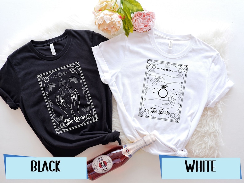 May include: Two t-shirts, one black and one white, with a graphic design of hands holding a ring and celestial symbols. The text on the black shirt reads 'The Groom' and the text on the white shirt reads 'The Bride.'