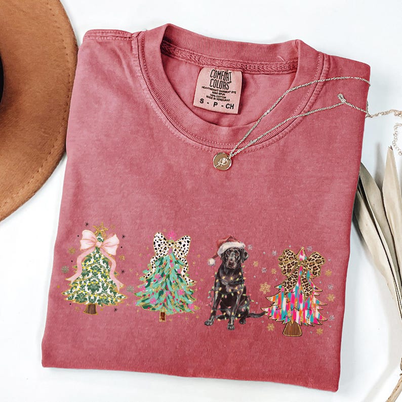 May include: A red t-shirt with a Christmas tree design featuring a black Labrador Retriever wearing a Santa hat. The trees are decorated with bows and ornaments. The shirt has the text "COMFORT COLORS" on the tag.