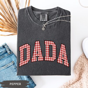 K&ouml;nnte beinhalten: Ein dunkelgraues T-Shirt mit dem Wort "DADA" in rot-wei&szlig;em Vichy-Druck. Das Shirt hat einen Rundhalsausschnitt und kurze &Auml;rmel. Eine silberne Halskette mit Anh&auml;nger ist auf dem Shirt drapiert. Das Wort "PEPPER" ist unten links aufgedruckt.