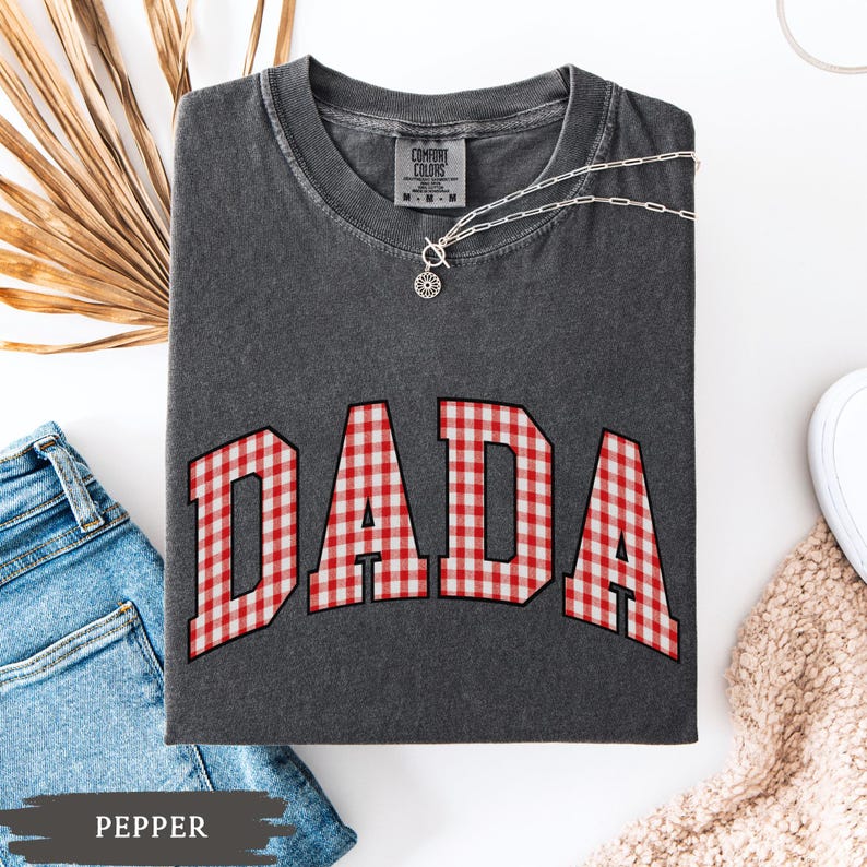 K&ouml;nnte beinhalten: Ein dunkelgraues T-Shirt mit dem Wort "DADA" in rot-wei&szlig;em Vichy-Druck. Das Shirt hat einen Rundhalsausschnitt und kurze &Auml;rmel. Eine silberne Halskette mit Anh&auml;nger ist auf dem Shirt drapiert. Das Wort "PEPPER" ist unten links aufgedruckt.